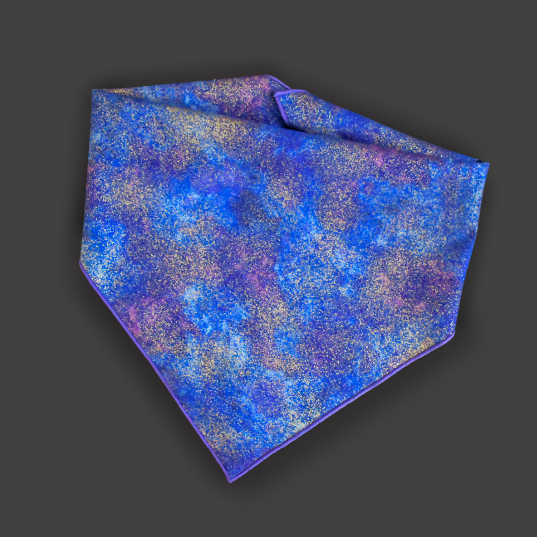 A flat lay of a bandana. A blue and purple cloudy print with flecks of metallic gold and a purple rolled hem on a dark gray backdrop.