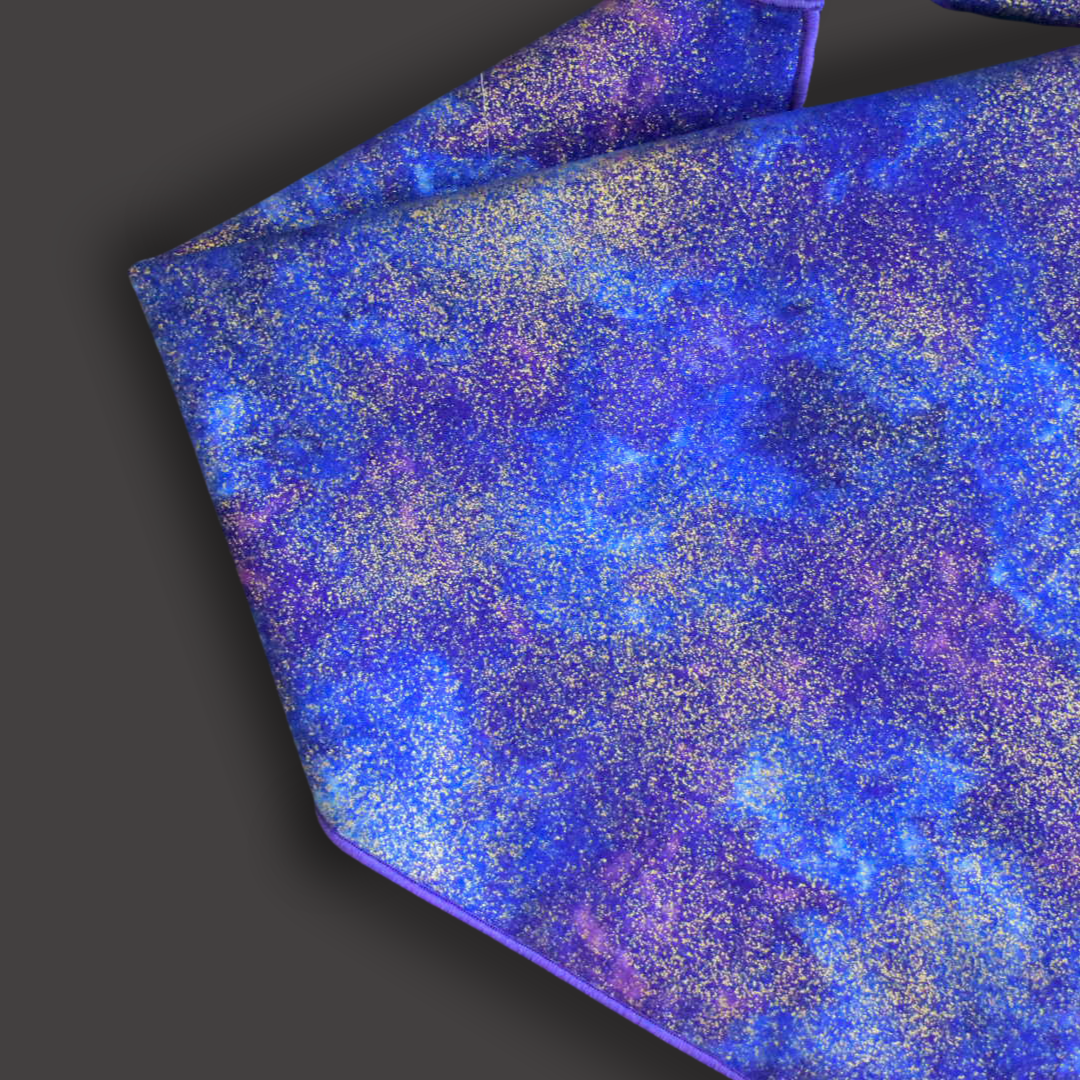 A zoomed in flat lay of the left 3/4 of a bandana. A blue and purple cloudy print with flecks of metallic gold and a purple rolled hem on a dark gray backdrop.