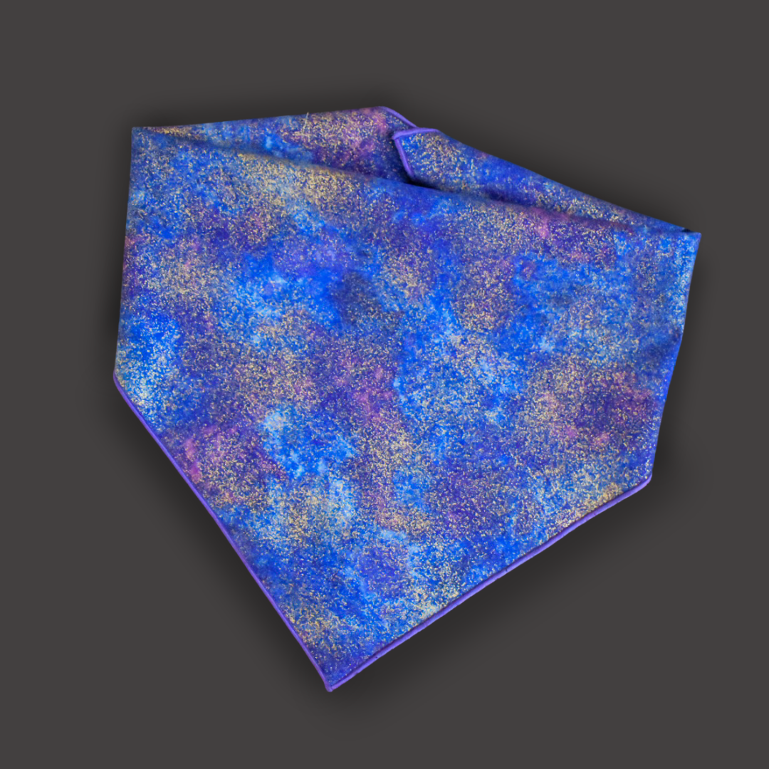 A flat lay of a bandana. A blue and purple cloudy print with flecks of metallic gold and a purple rolled hem on a dark gray backdrop. 