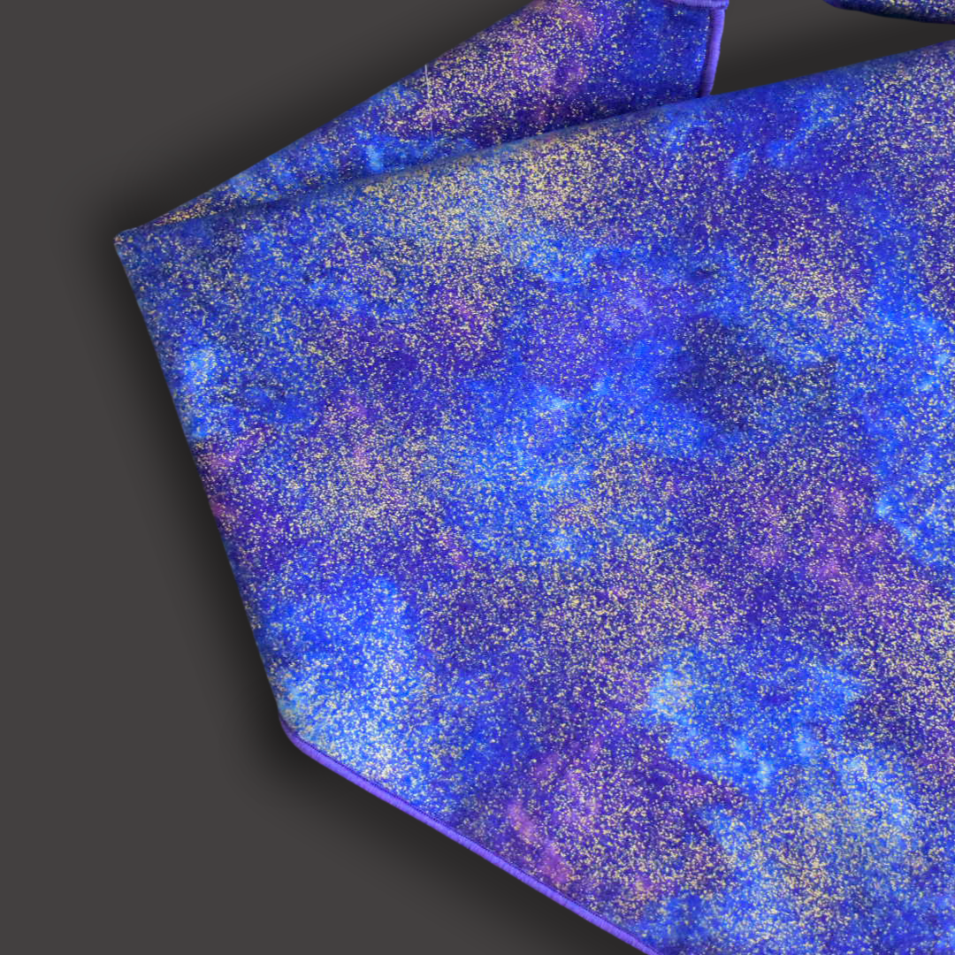 A zoomed in flat lay of the left 3/4 of a bandana. A blue and purple cloudy print with flecks of metallic gold and a purple rolled hem on a dark gray backdrop. 